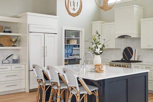 Two rattan textured pendants hang above a Welborn Bleu Expanded Island with chrome accents. White Cabinetry on the perimeter allows the space to feel airy and light while the navy island grounds the space.