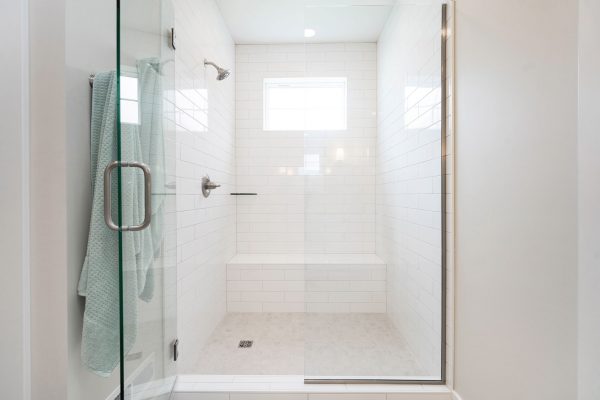 Walk-in shower with a glass enclosure & built-in bench along the widest wall. Clear glass and gold bath accessories catch the natural light streaming in from the clerestory window high above the bench wall, illuminating the white marbled tile along the floor and walls.